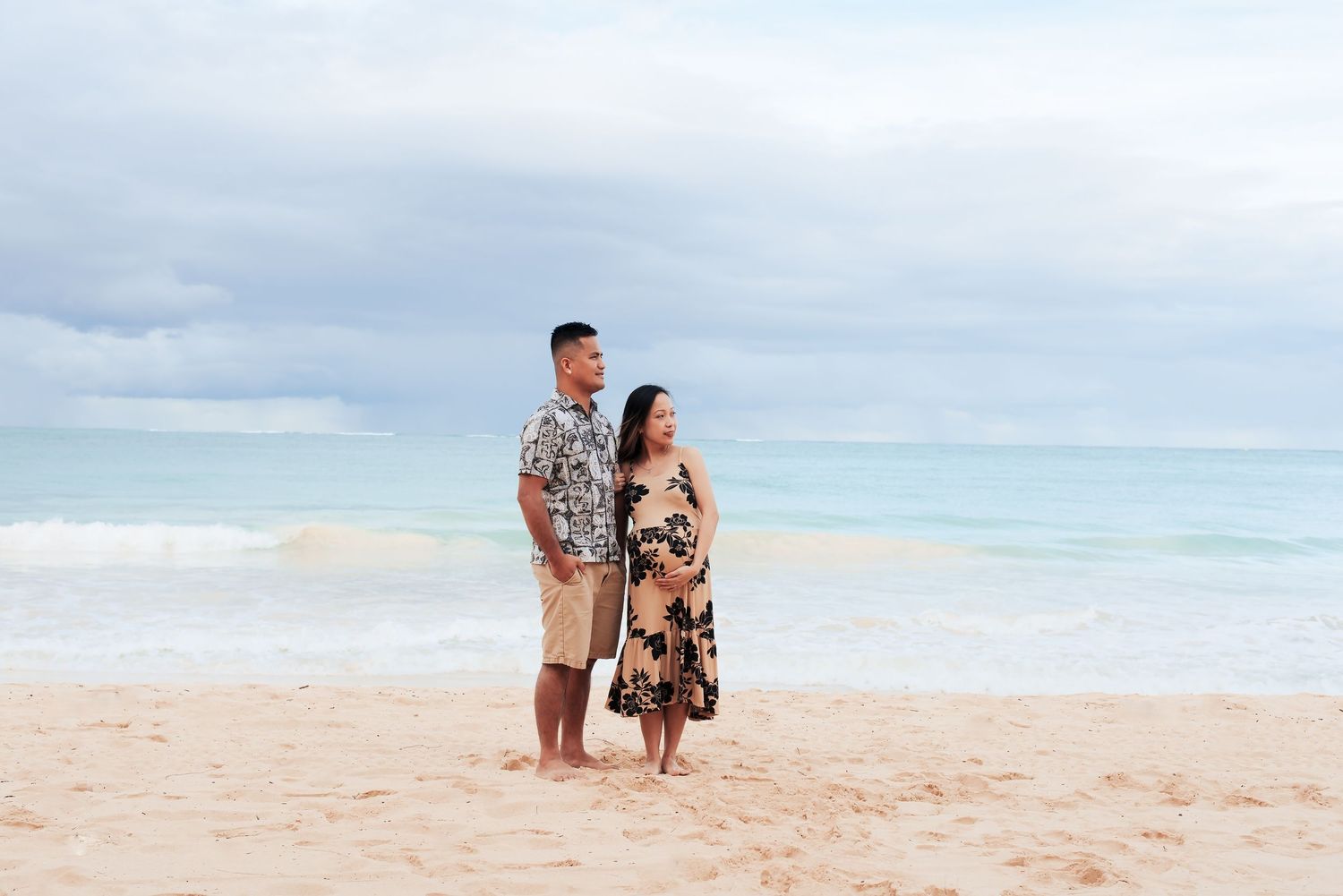 Couple standing on a sandy beach in tropical outfits, smiling at each other with turquoise ocean waves in the background.