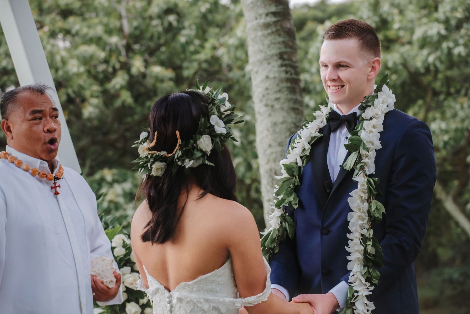Groom in navy suit with floral lei smiles at bride during outdoor Hawaiian wedding ceremony with officiant.