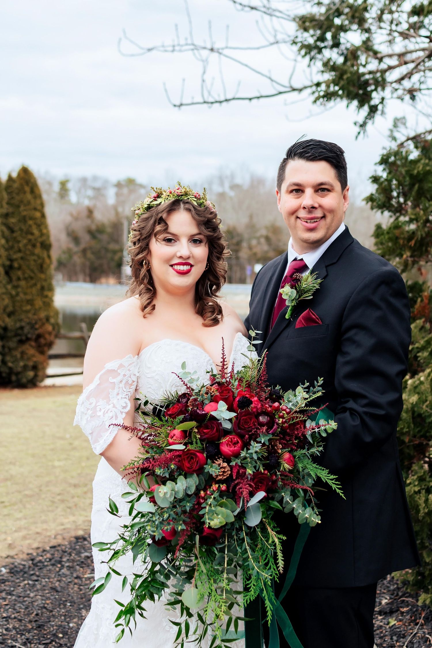 Winter wedding couple poses outdoors with a dramatic burgundy and green floral bouquet near evergreen trees.