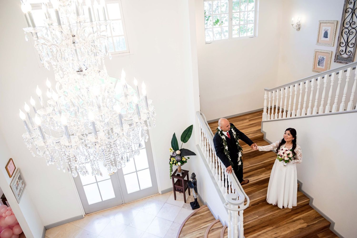 Bride and groom descend elegant staircase beneath crystal chandelier in bright white wedding venue.