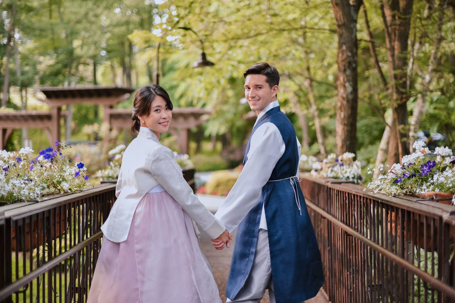A couple wearing traditional Korean hanbok outfits holds hands while standing on a wooden bridge in a garden setting.