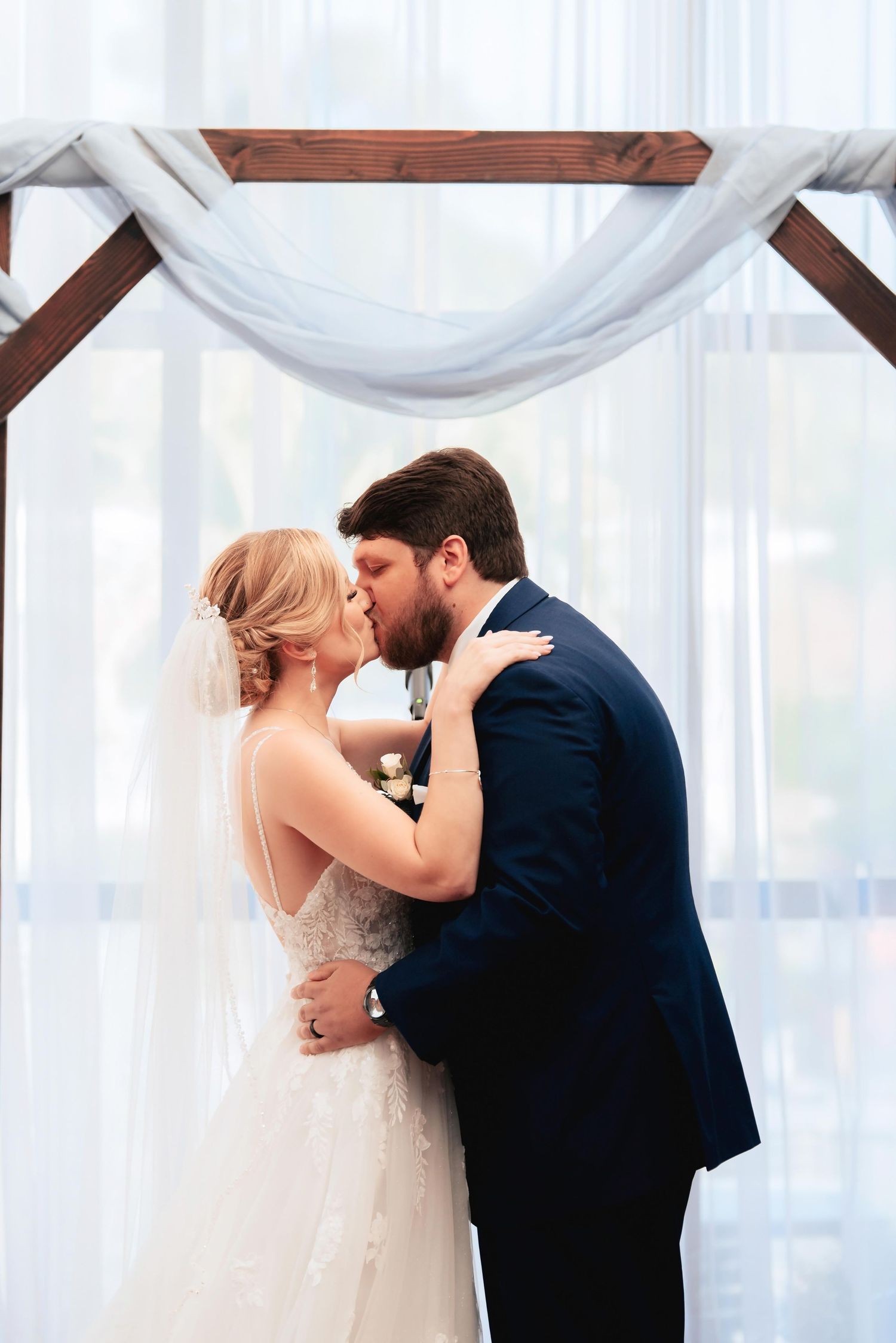 A newly married couple shares a romantic kiss under white flowing fabric draping at their wedding ceremony.