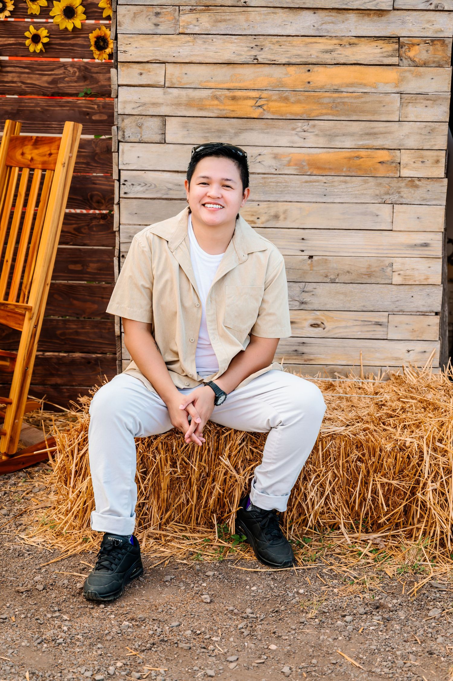 Young man smiling, sitting on a hay bale near a rustic wooden wall with sunflowers, wearing beige shirt and white pants.