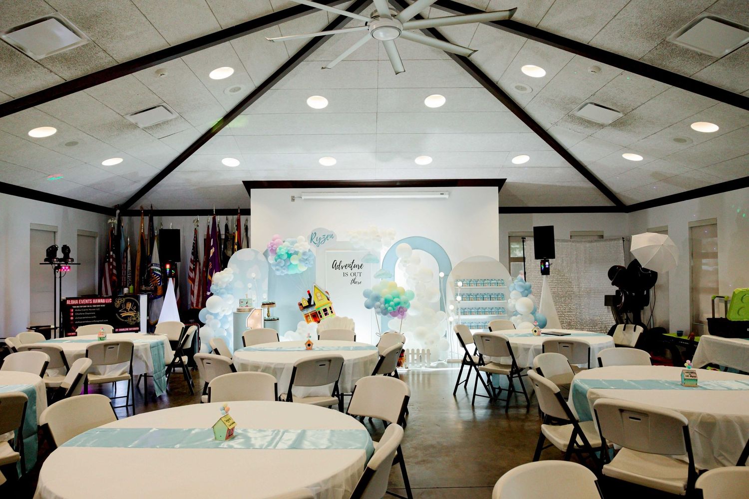 Elegant baby shower venue with round tables, teal decor, balloon arch, and a decorative backdrop in a banquet hall.