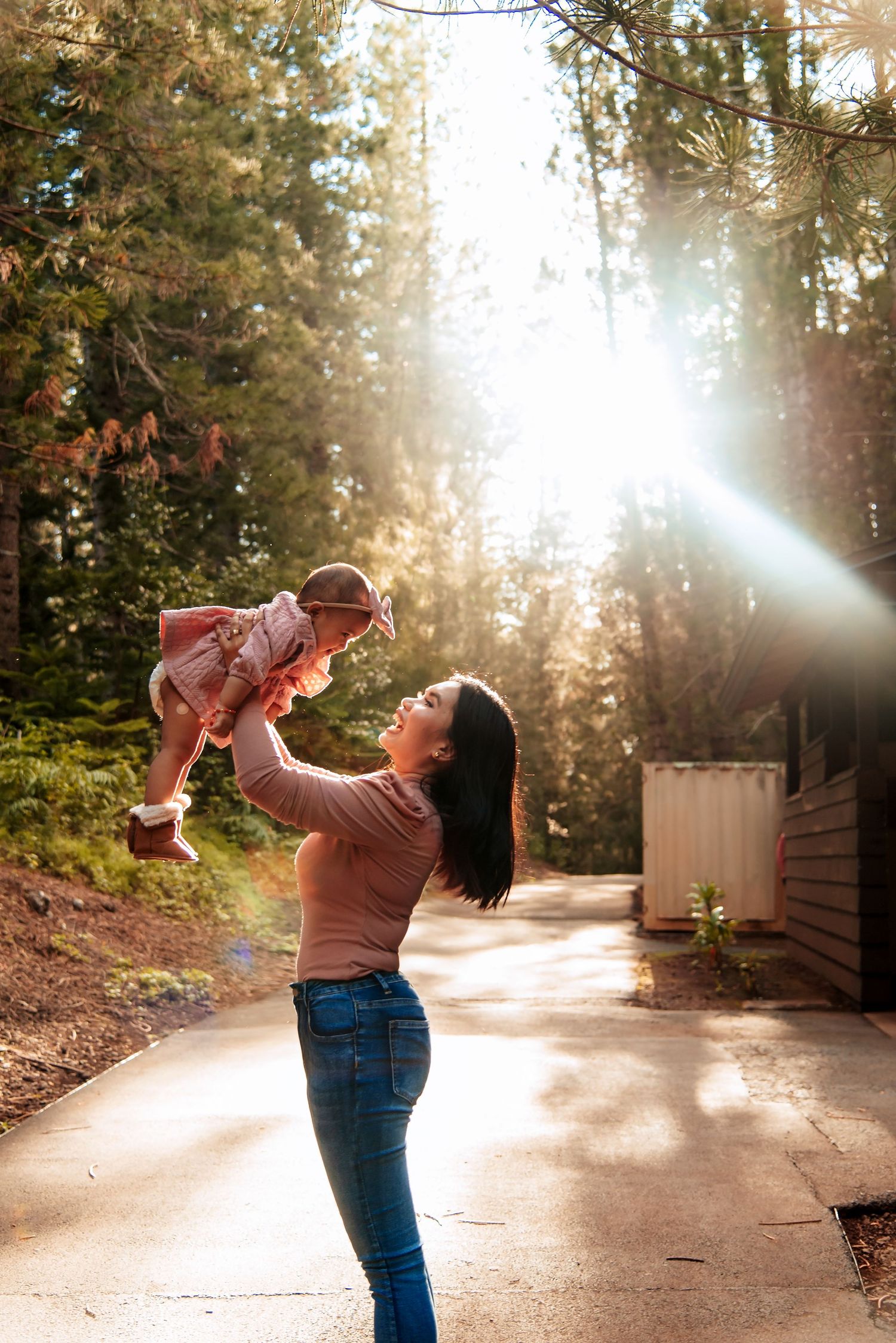 A mother joyfully lifts her baby in the air outdoors, surrounded by tall pine trees with sunlight streaming through.