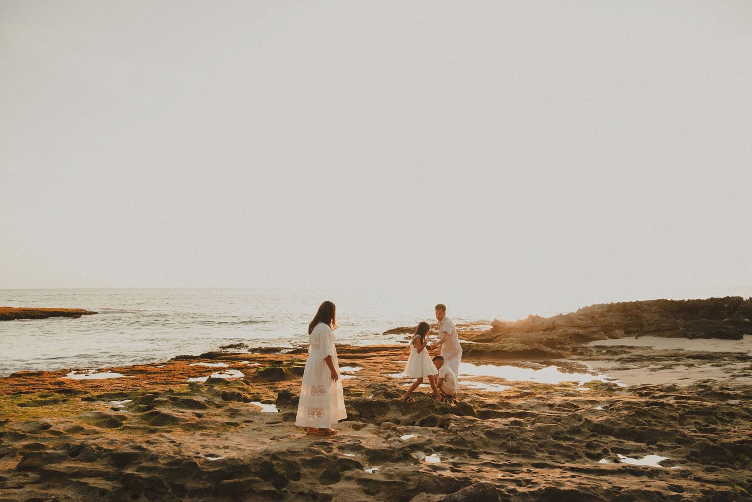 Family in white outfits playing on rocky beach at golden sunset near ocean waves.