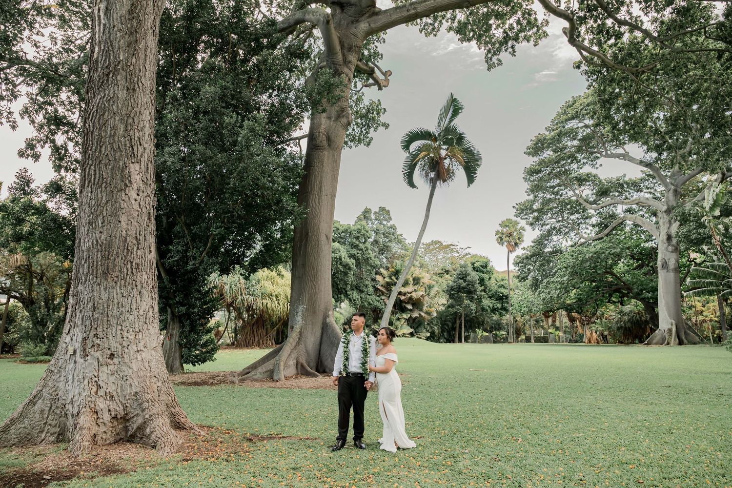 Couple stands in lush tropical garden with towering trees and palm trees on their wedding day.