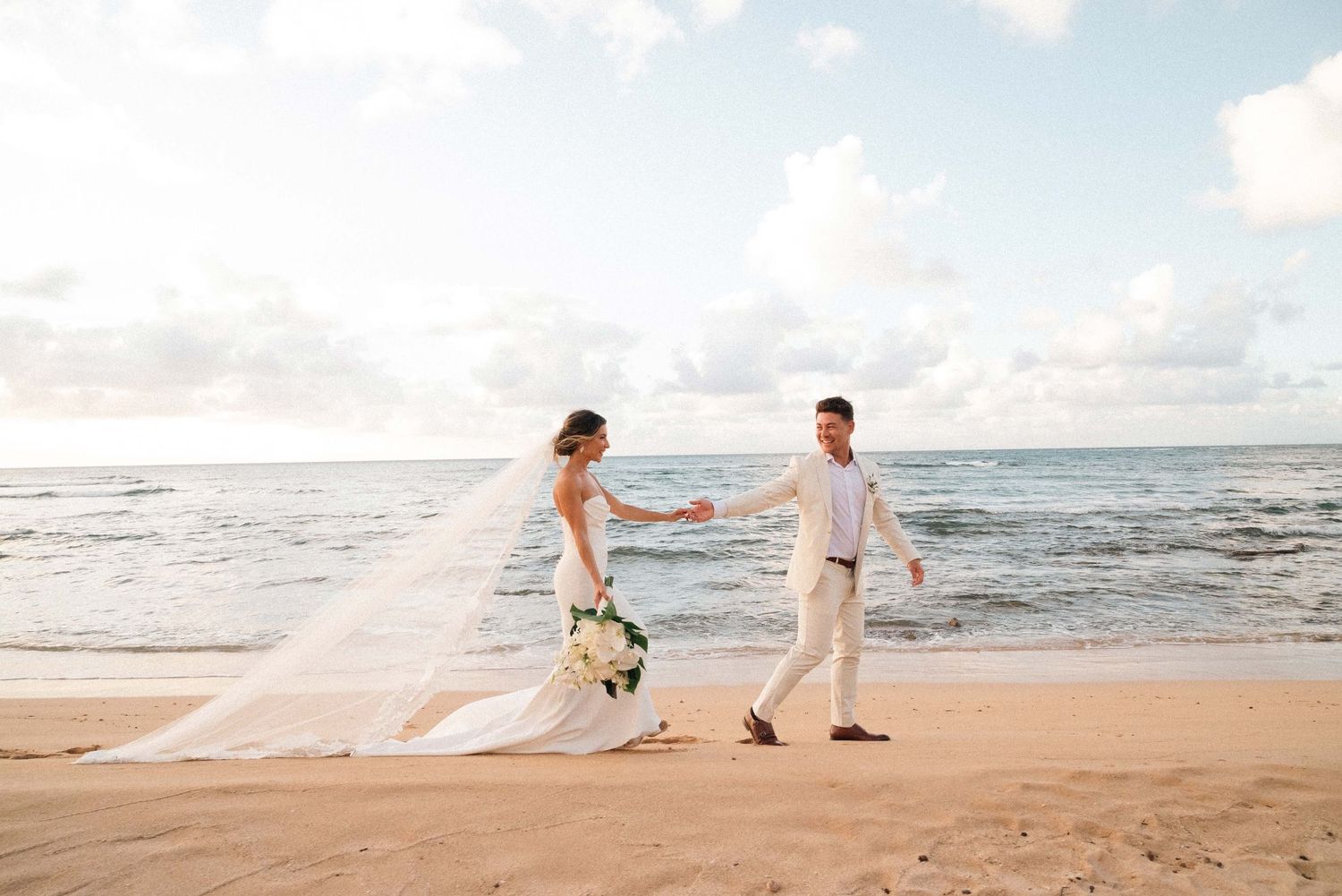 Bride and groom holding hands walking on a sandy beach with ocean waves, wedding veil flowing in the breeze.