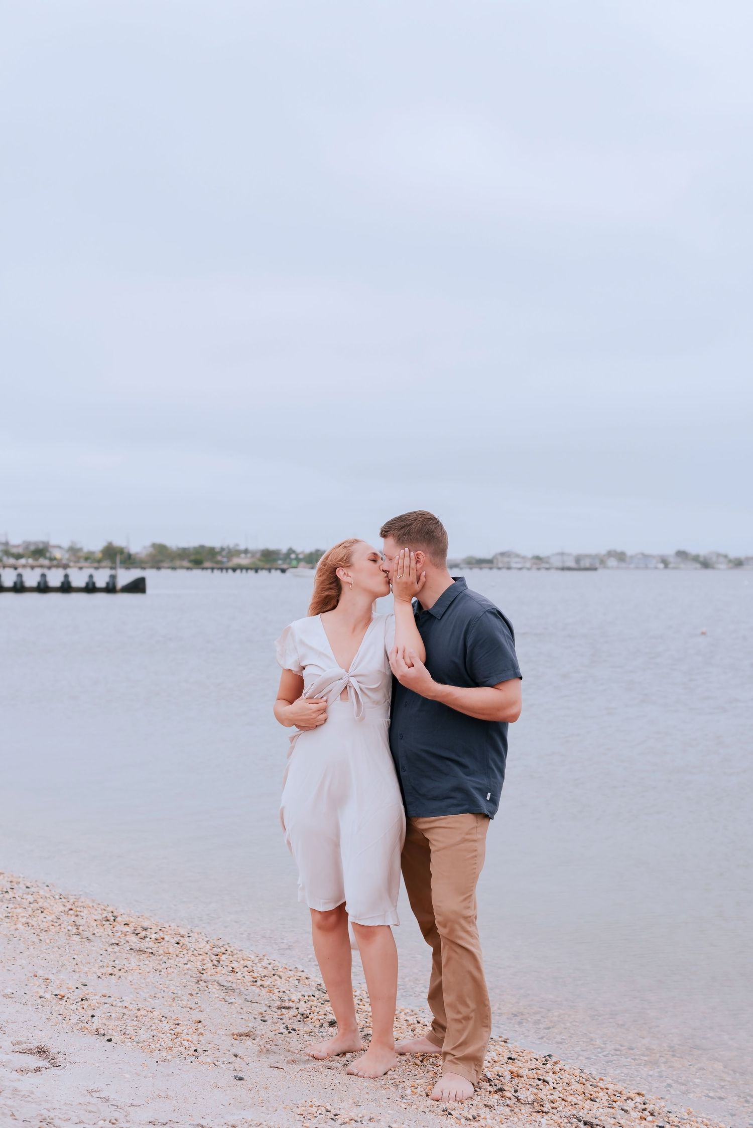 A couple shares a romantic kiss on a sandy beach shore with calm water and cloudy skies in the background.