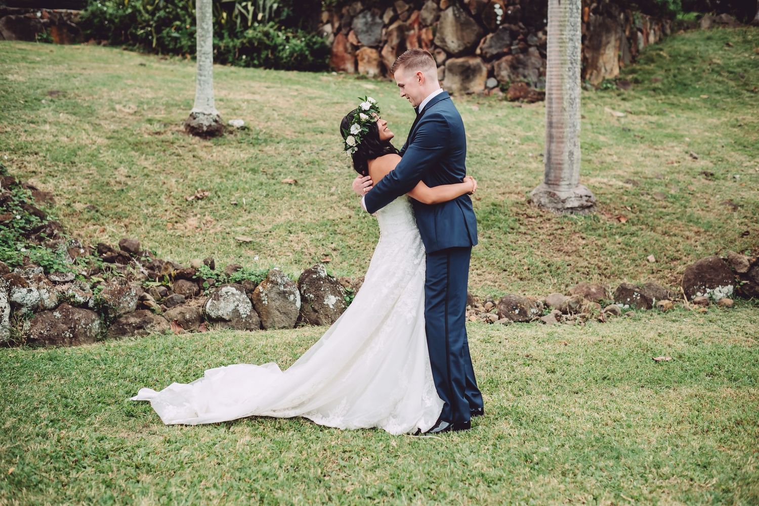 Bride and groom share a romantic embrace on green lawn, bride in elegant white gown with train, groom in navy suit.