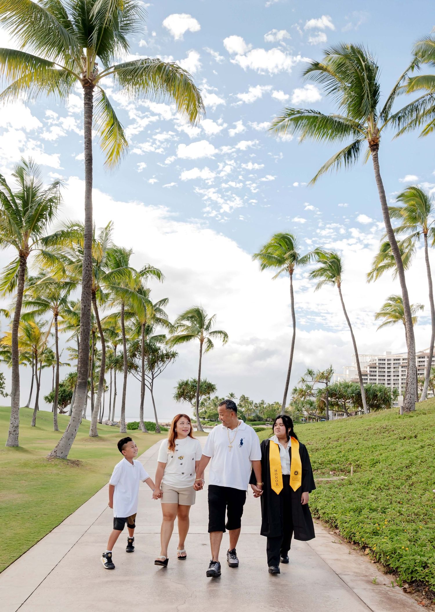 Family of four walks along a palm-lined path in Hawaii, with a graduate in cap and gown holding hands with loved ones.