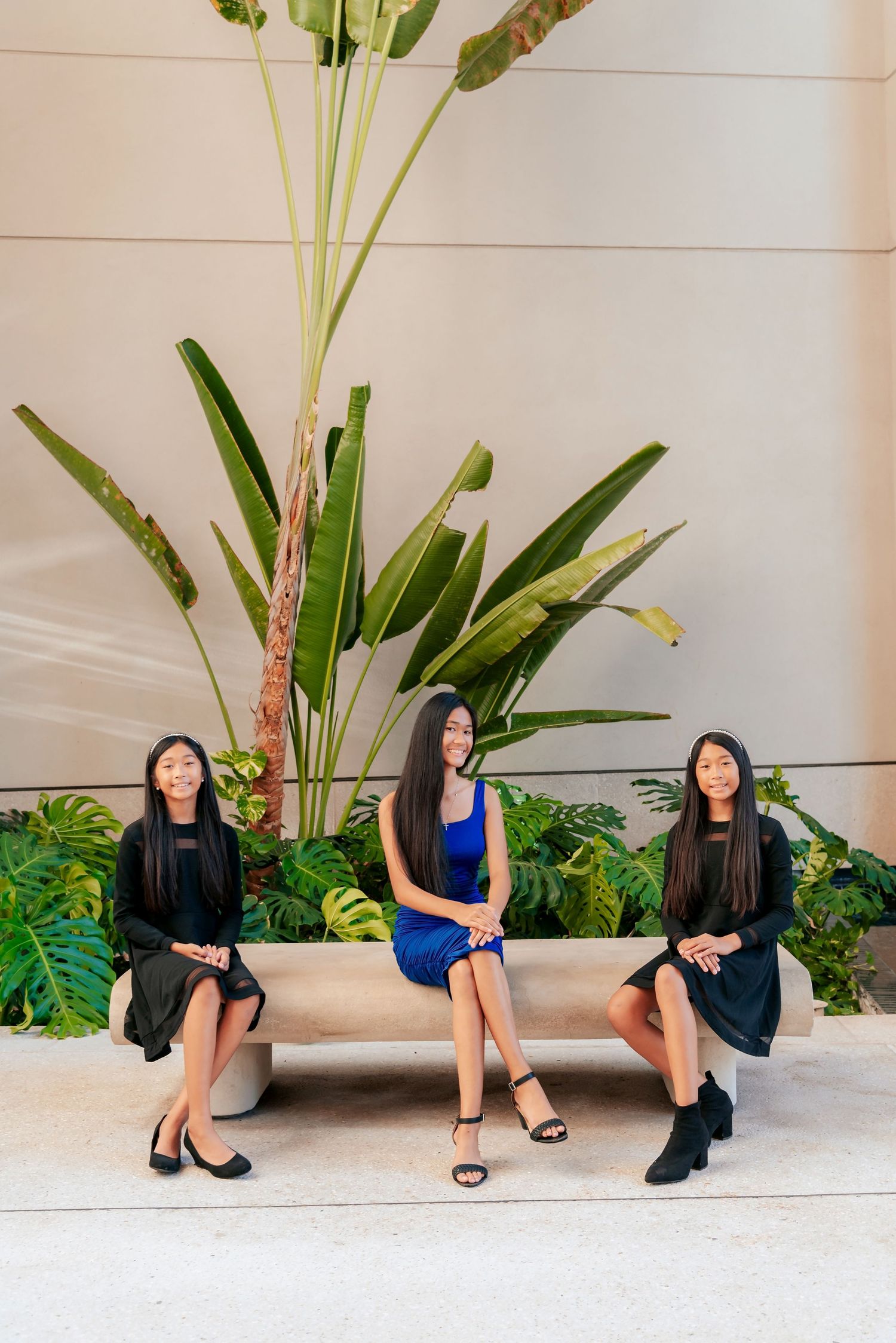 Three young women in elegant dresses sit on a stone bench surrounded by tropical plants.