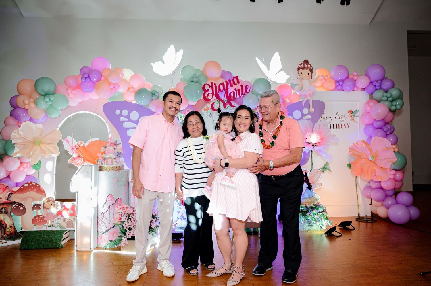 Family posing together at a colorful birthday party with balloon arch and floral decorations spelling Eliana Marie.