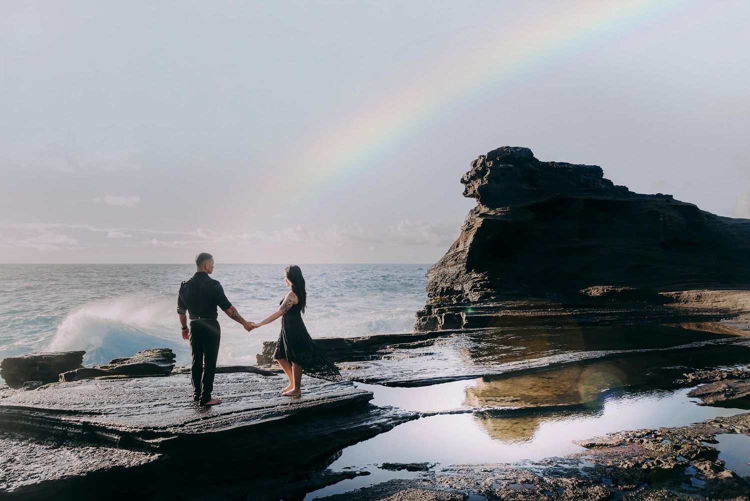 Couple holding hands on rocky shore with rainbow over ocean waves.