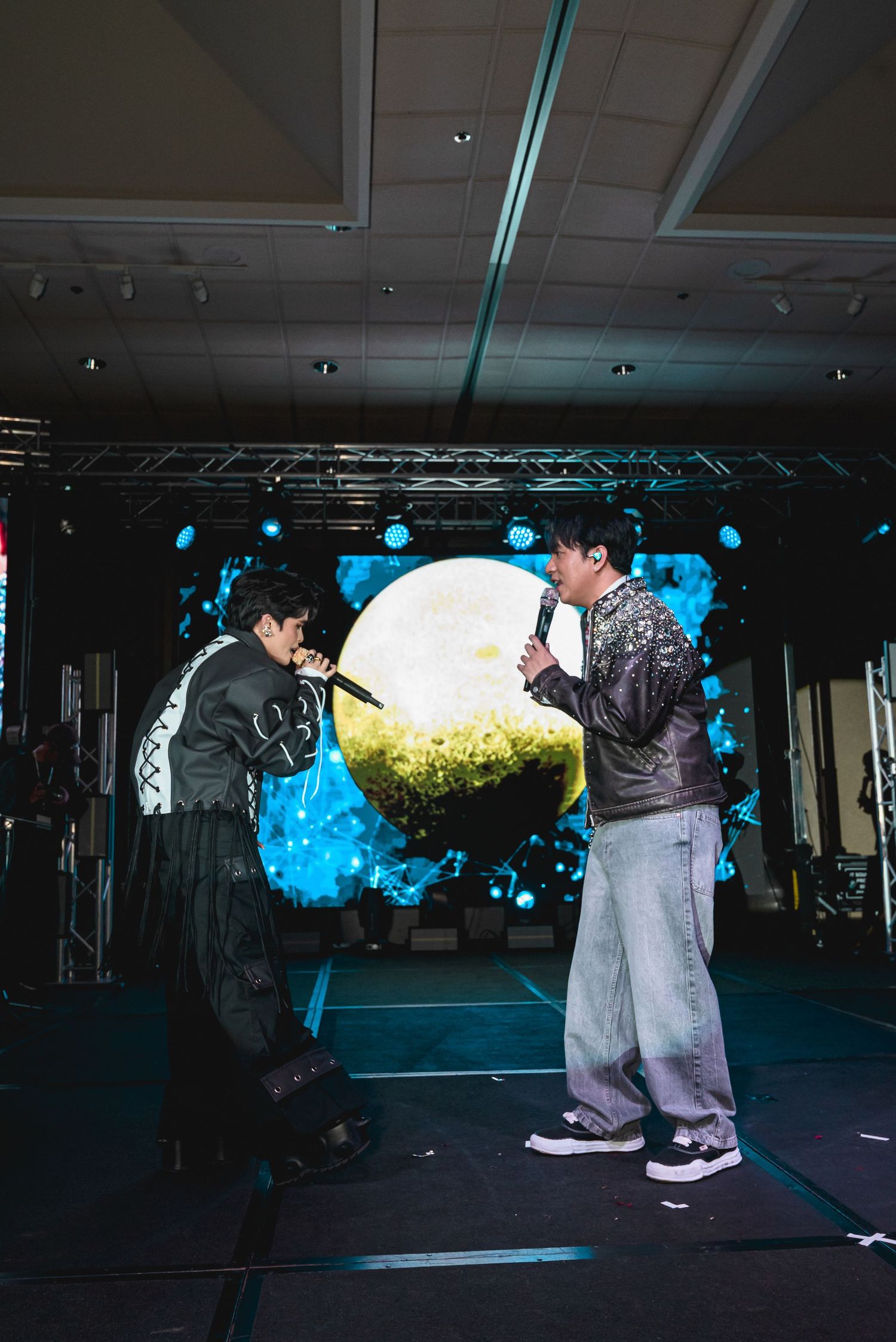 Two performers singing on stage with a glowing moon backdrop and blue light effects at a live concert event.