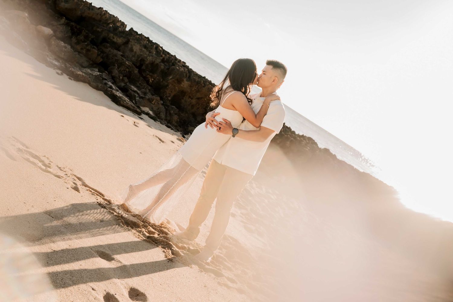 Couple embracing romantically on a sunny sandy beach near rocky cliffs, dressed in white outfits.