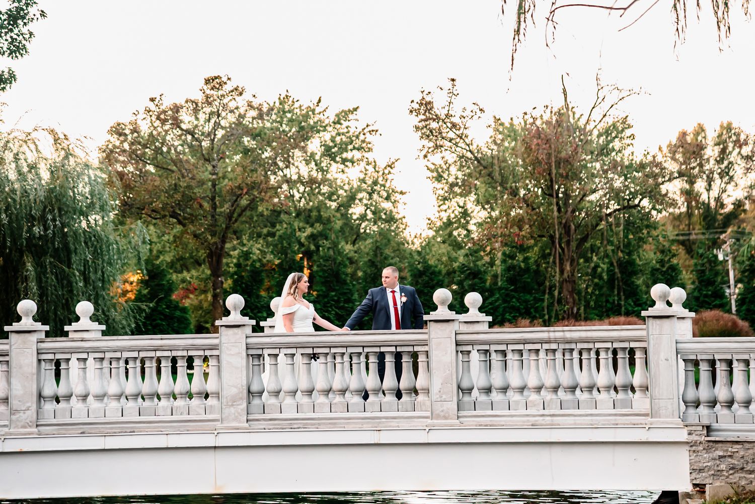 Wedding party stands along elegant white balustrade railing at sunset during outdoor garden ceremony.