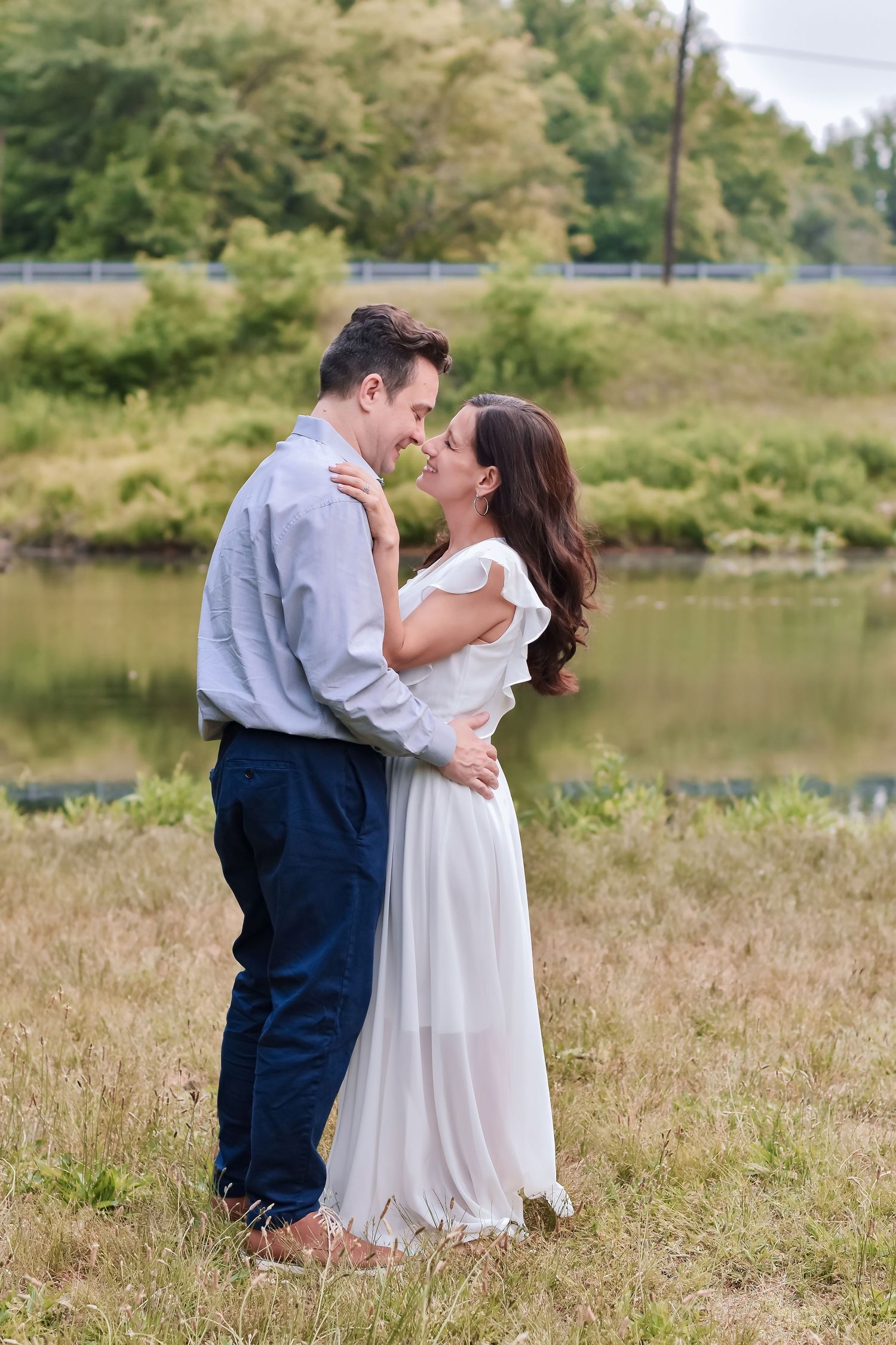Romantic couple embracing outdoors near a serene pond, woman in white dress and man in blue shirt, surrounded by lush greenery.