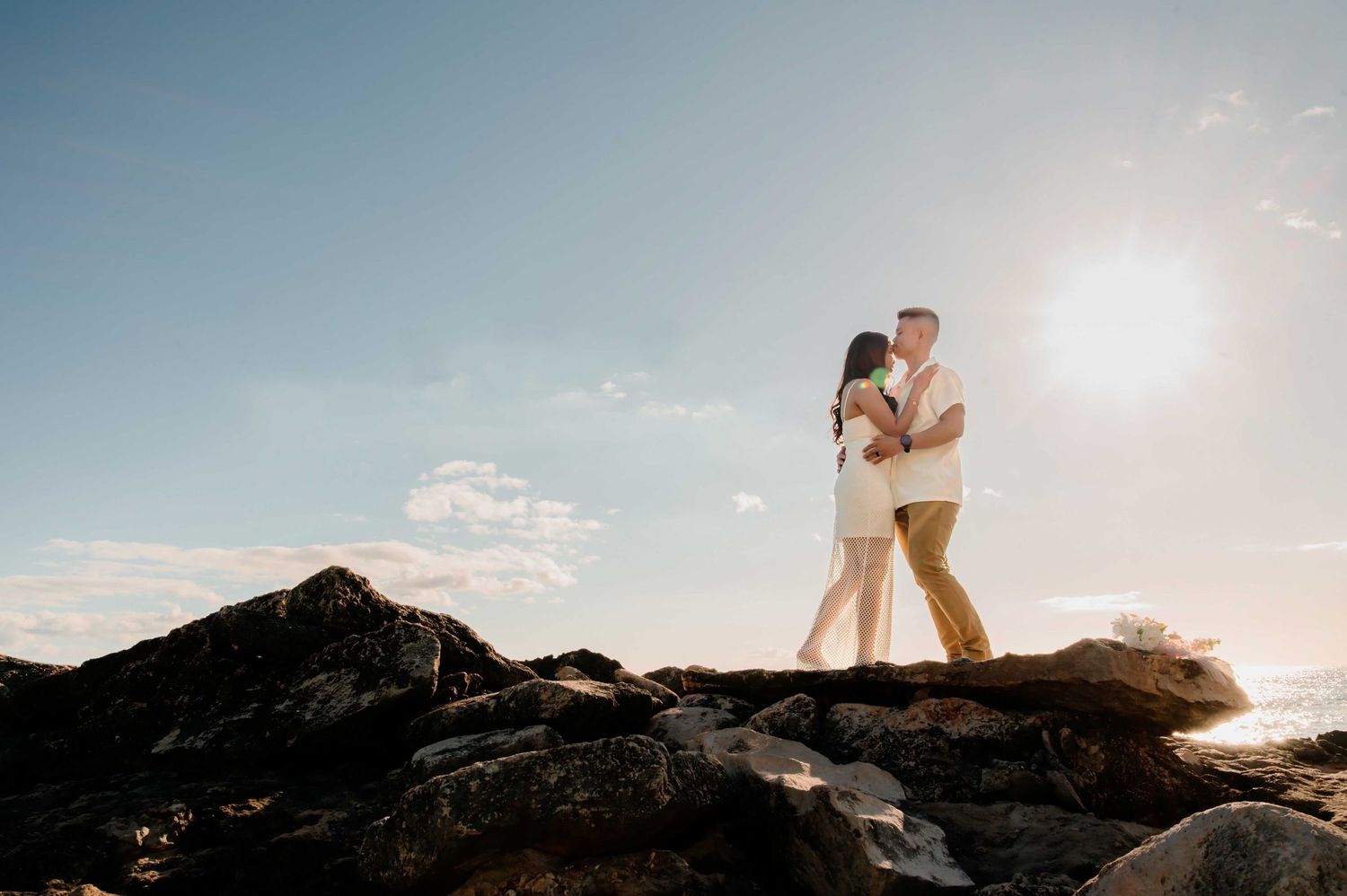 Couple embracing on rocky shoreline at golden hour with bright sun and blue sky backdrop.