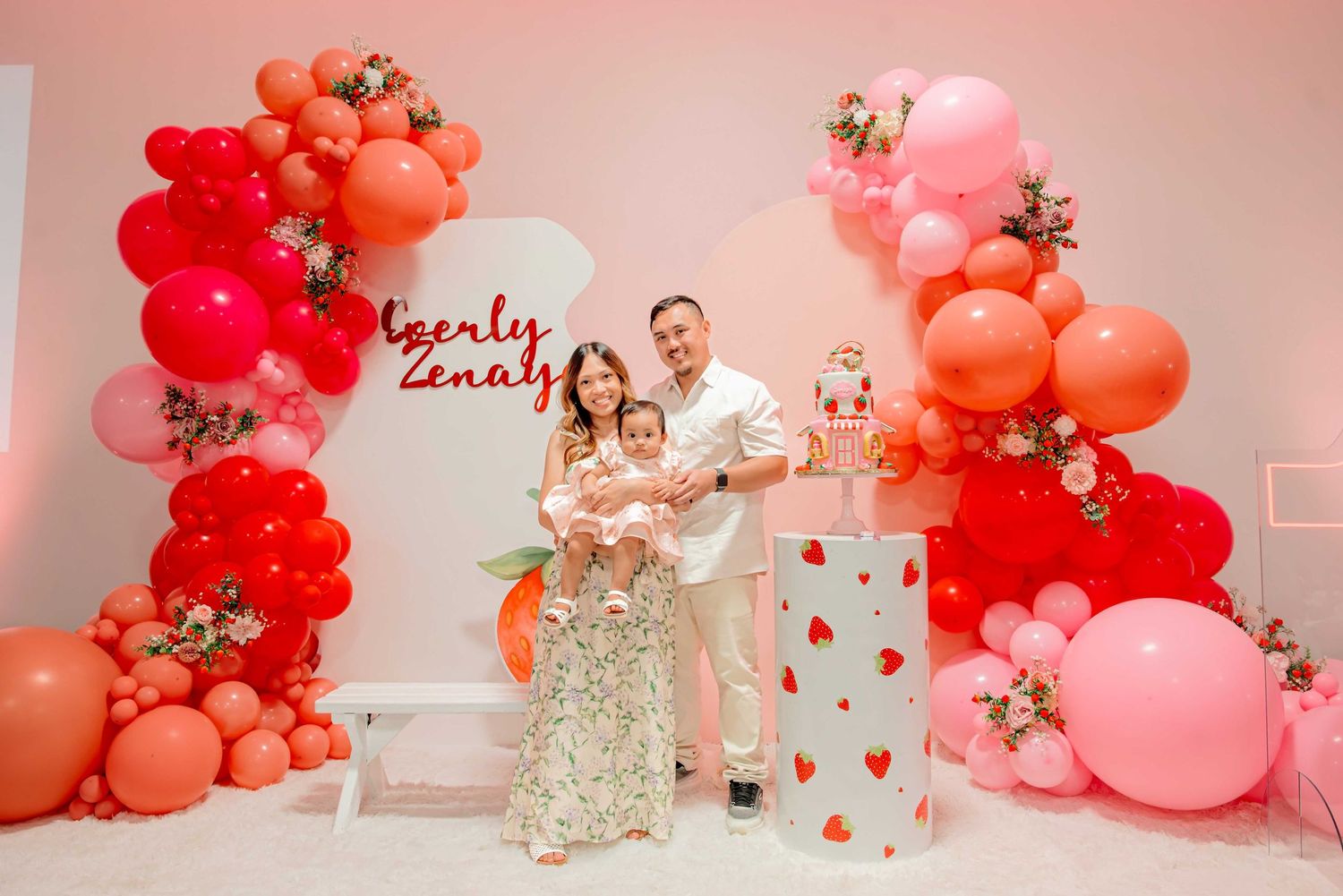 Family posing in front of colorful balloon arch with pink red and orange balloons at a birthday party celebration.