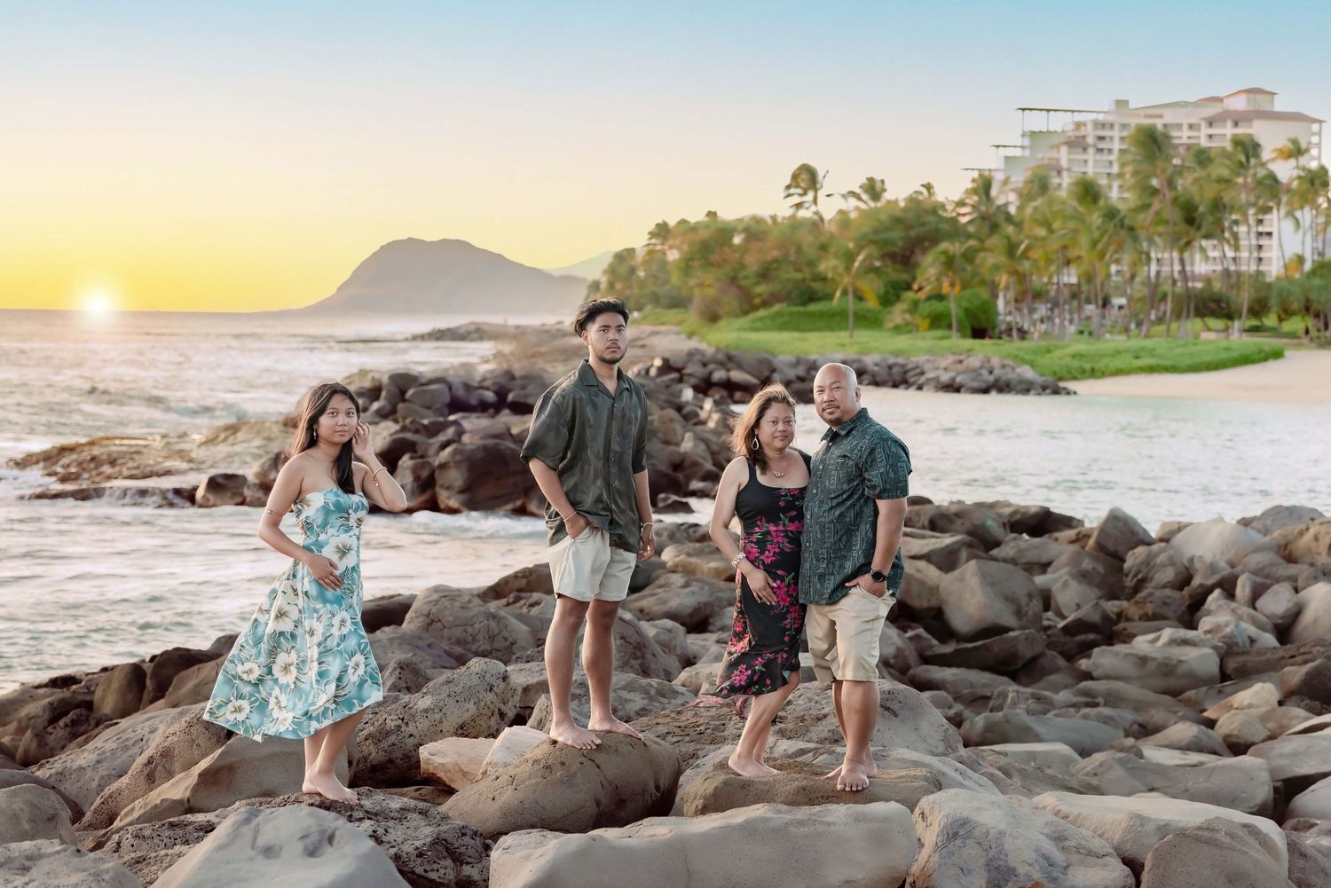 Family of four posing on rocky Hawaiian beach at sunset with resort and palm trees in background.