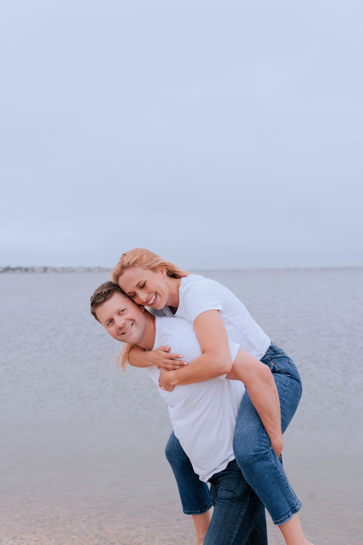 A happy couple laughing at the beach, man giving woman a piggyback ride, both wearing white shirts and jeans.