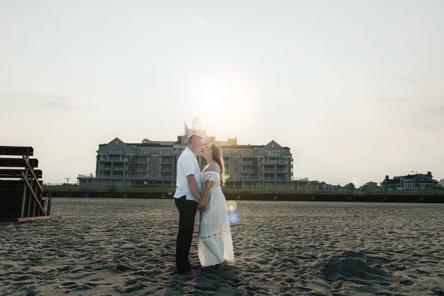 Couple sharing a romantic sunset kiss on a sandy beach with a resort building glowing in the golden light behind them.