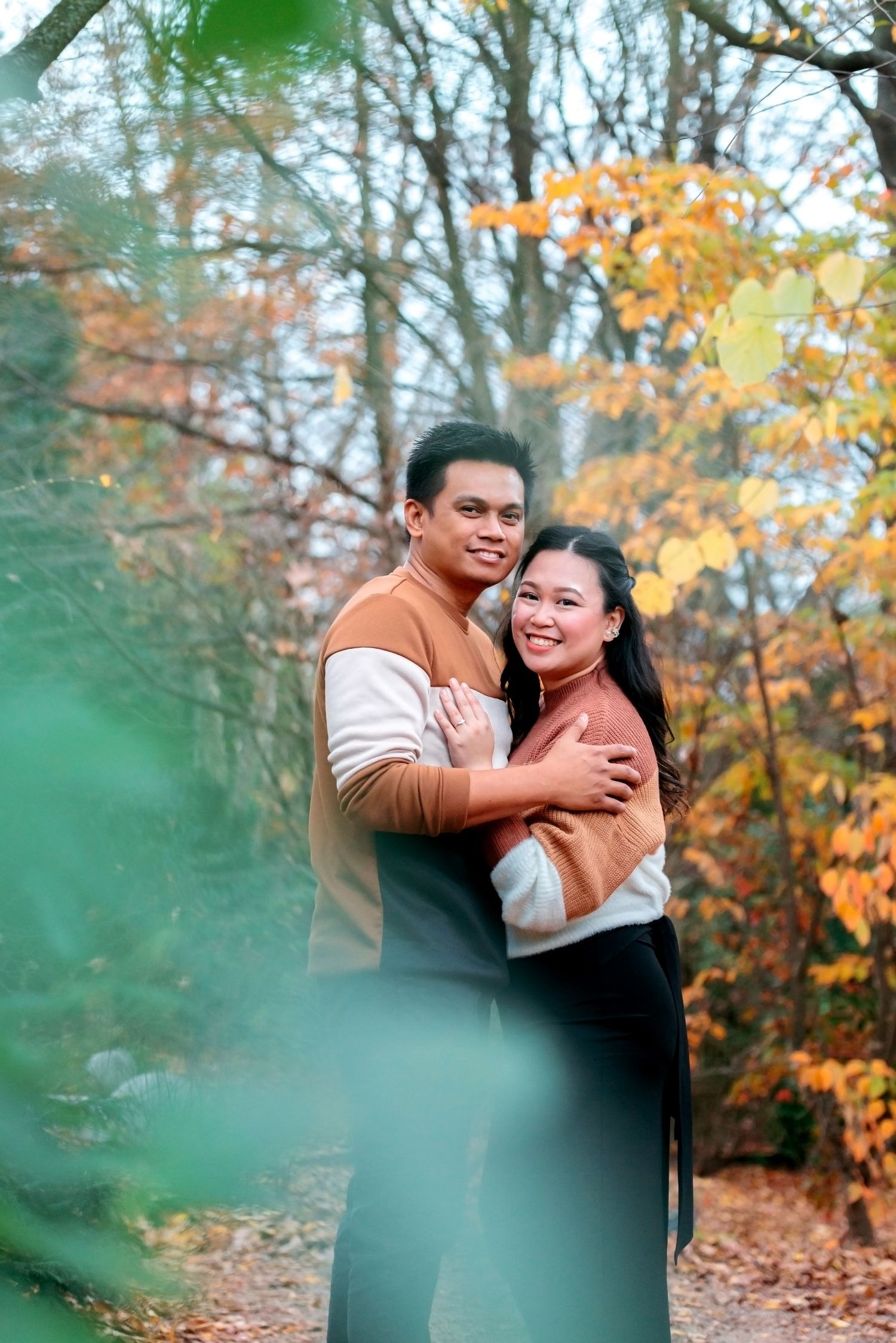 Two figures embrace lovingly amidst autumn foliage in a romantic outdoor photoshoot at the park.
