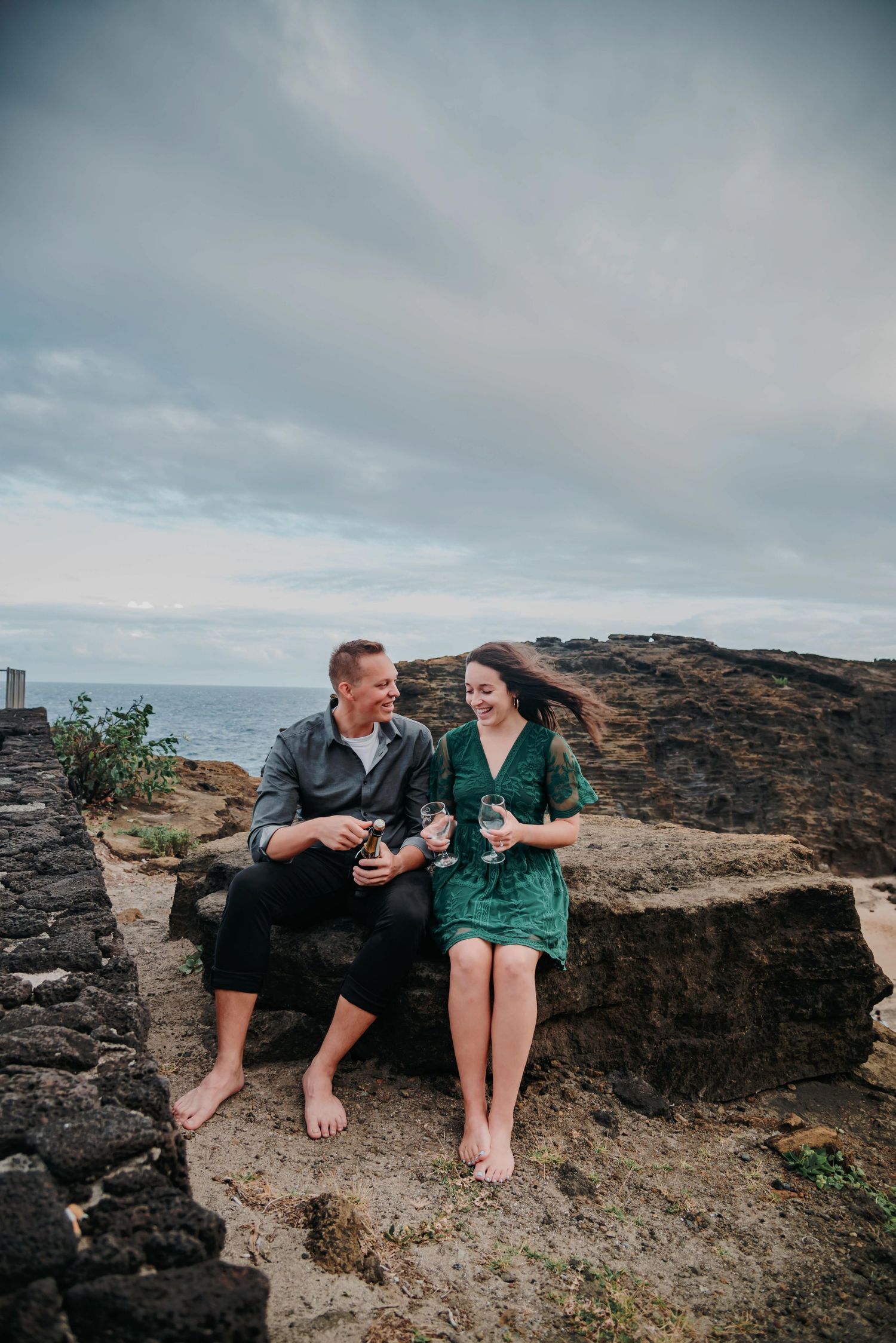 A couple enjoying drinks while sitting barefoot on coastal rocks under cloudy skies at sunset.