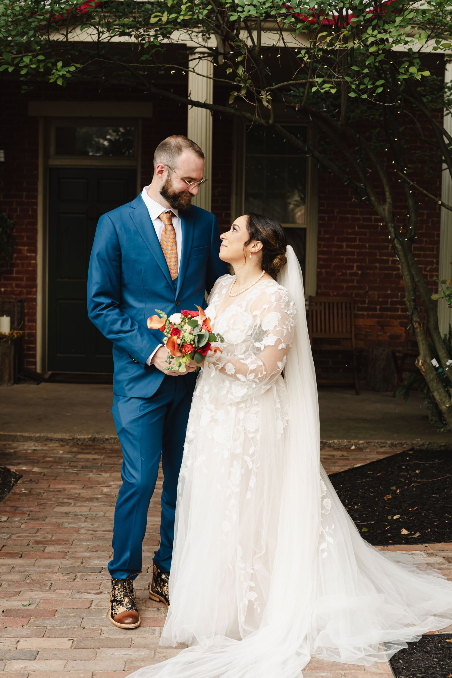 Couple shares intimate moment in wedding attire standing on brick patio with bride in lace gown and groom in blue suit.