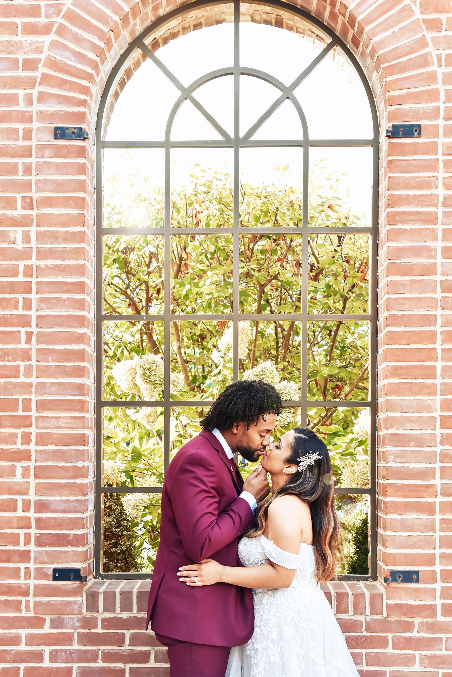 Couple shares romantic moment by arched brick window with garden view in burgundy suit and white dress.