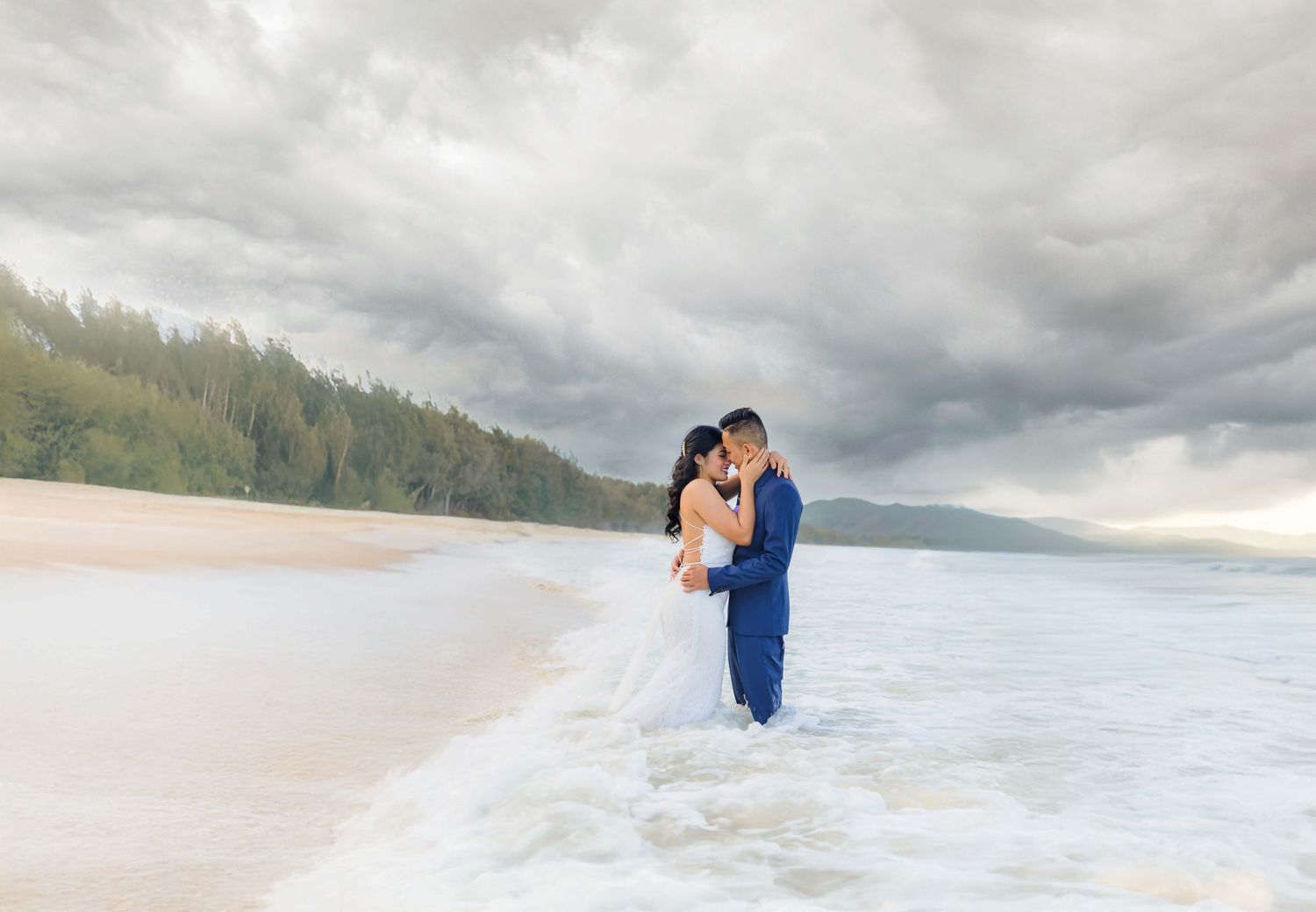 Couple in wedding attire embracing on a tropical beach with dramatic stormy skies and ocean waves.