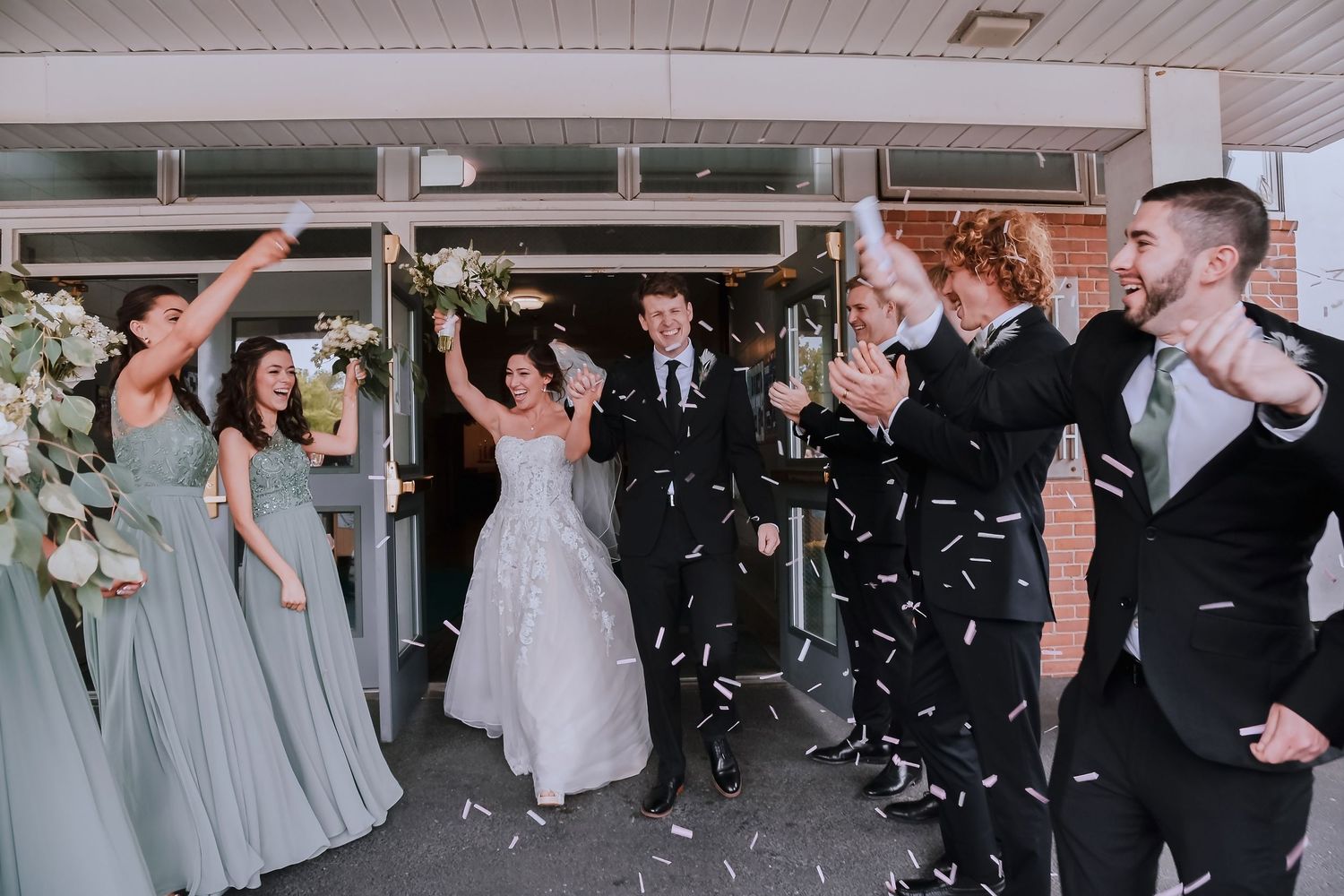 Bride and groom exit wedding venue surrounded by bridesmaids and groomsmen celebrating with confetti cannons.