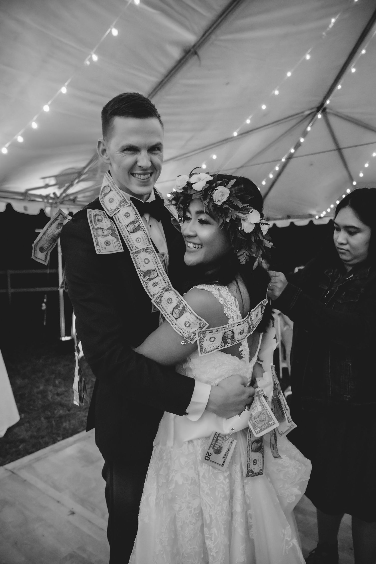 Wedding guests playfully shower the couple with dollar bills during a celebratory dance under a white tent with string lights.