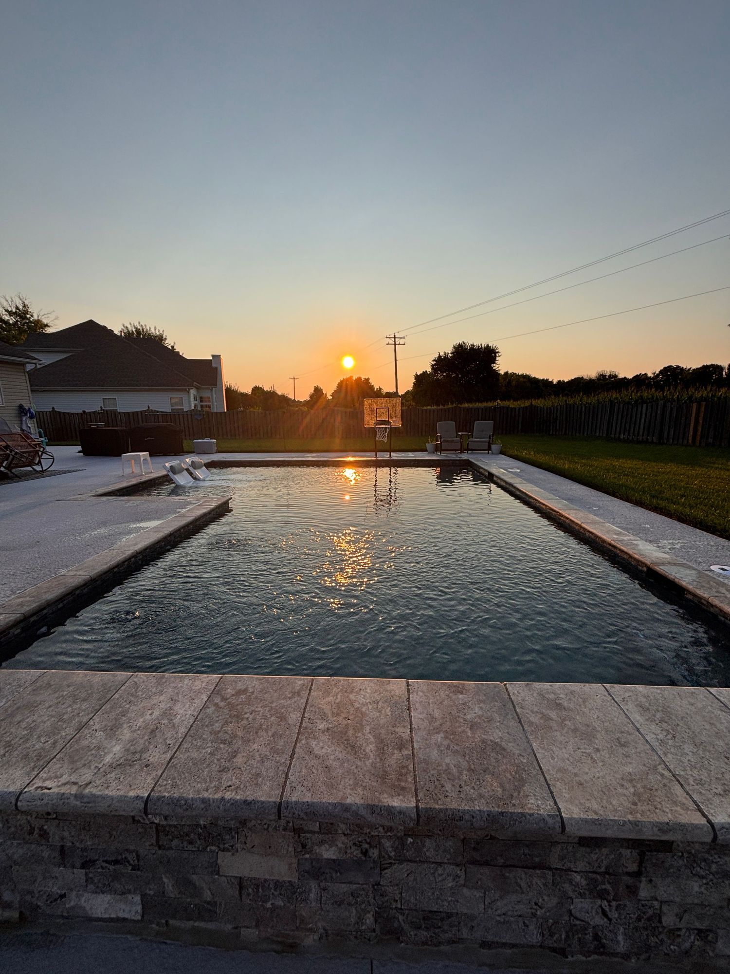 Stunning rectangular backyard pool with travertine coping reflecting a golden sunset in a suburban yard.