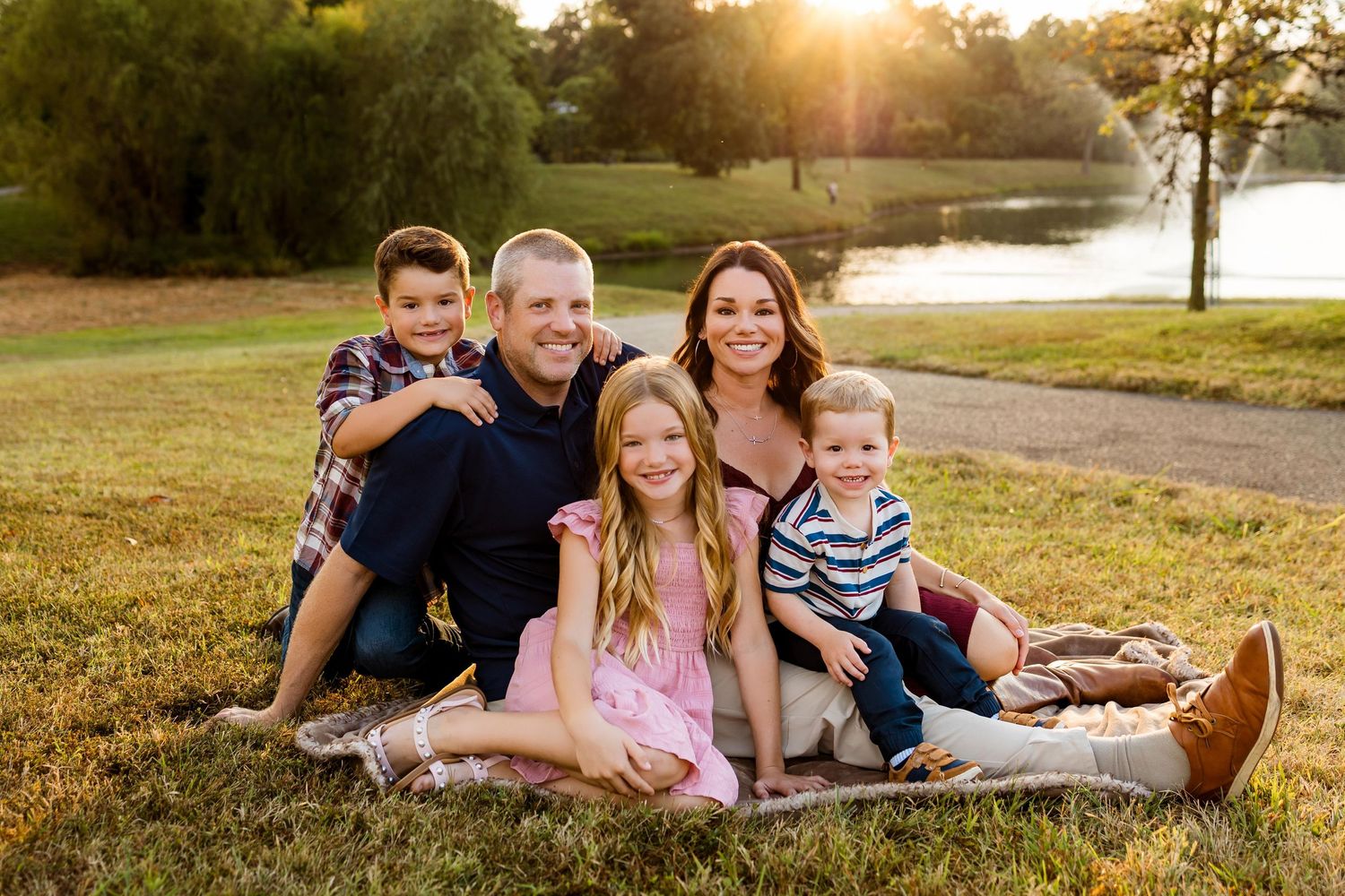 A family relaxes together on grassy ground during golden hour sunset in a park setting.