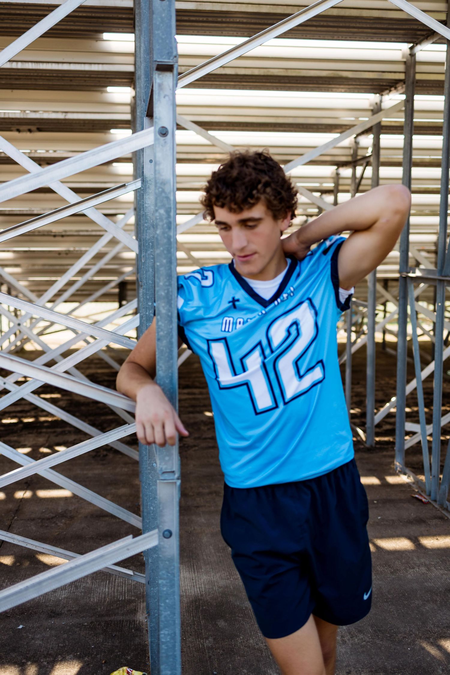Young athlete in light blue jersey and navy shorts stretches against metal bleacher supports at sports facility.