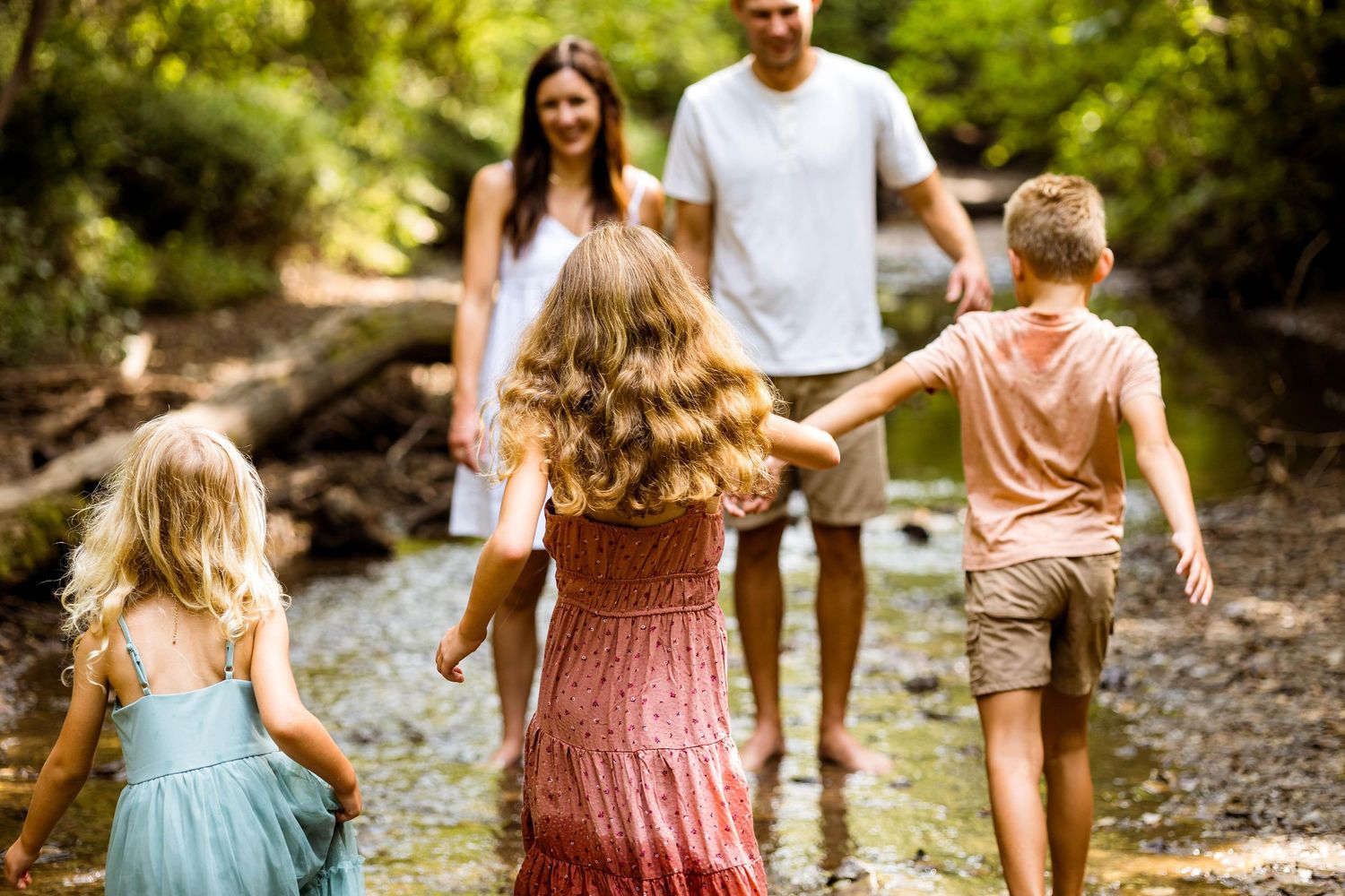 Family walking and playing together along a creek bed in sunny nature setting during summer outdoor adventure.