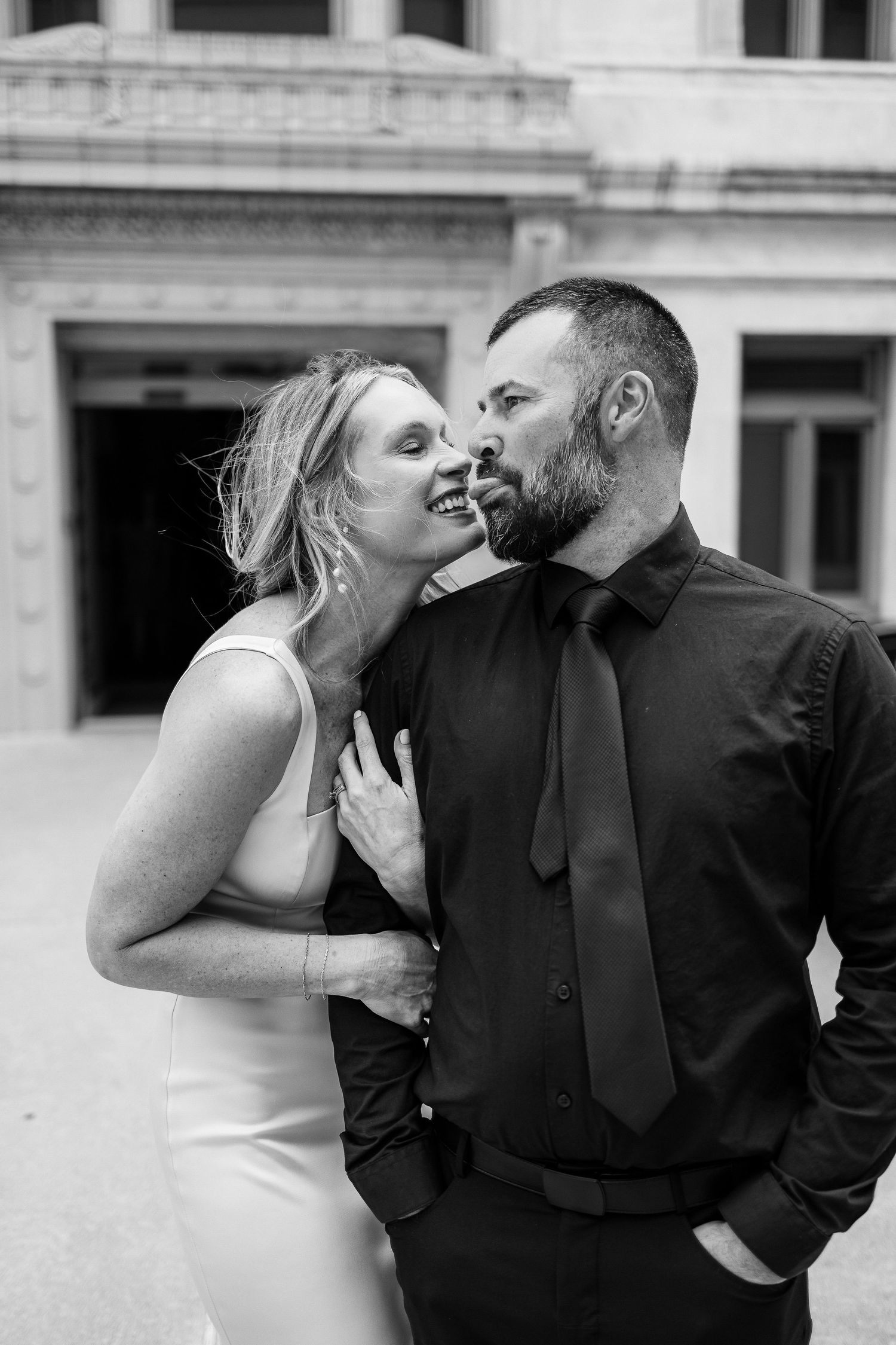 Black and white photo of stylish couple sharing intimate moment against classical architecture backdrop.