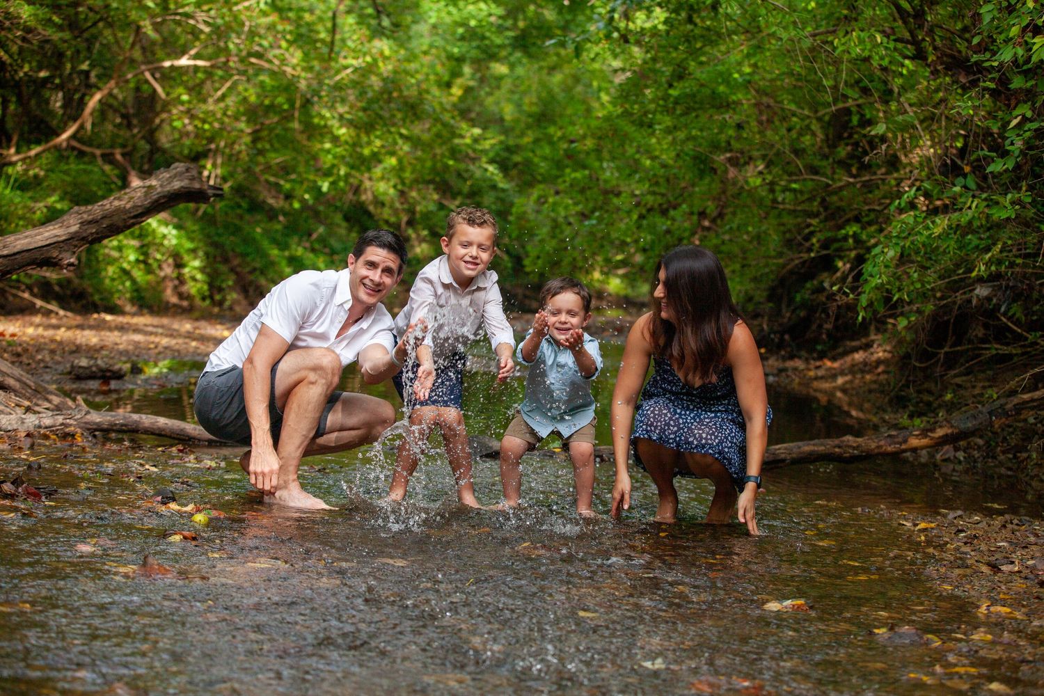 Group of friends crouching by a creek splashing in water surrounded by lush green forest on a summer day.