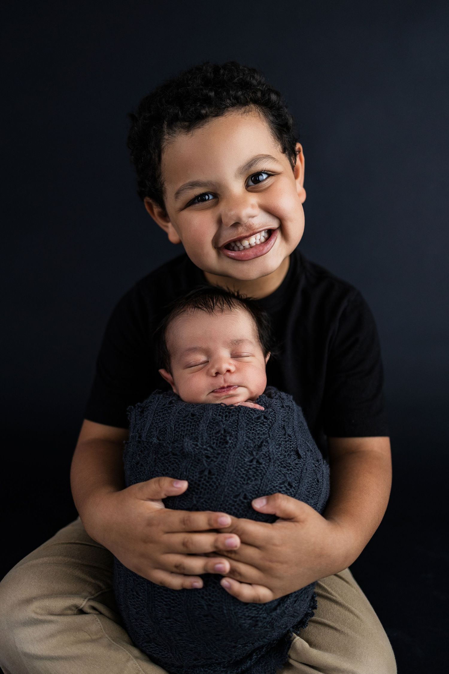 A joyful child holds a sleeping newborn wrapped in a dark blue knit blanket against a black background.
