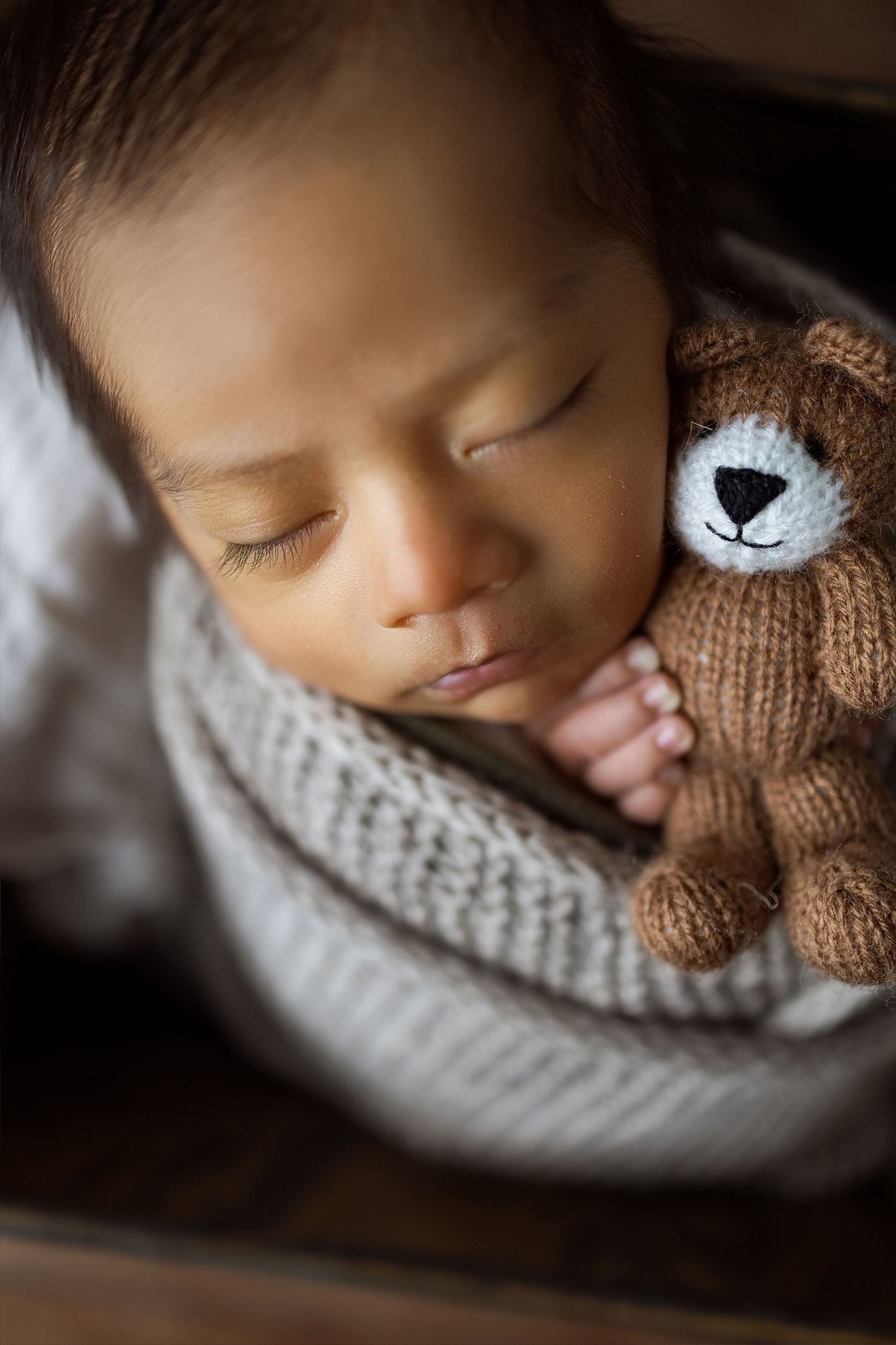 A sleeping newborn nestled in a cozy knit blanket with a plush teddy bear companion in soft lighting.