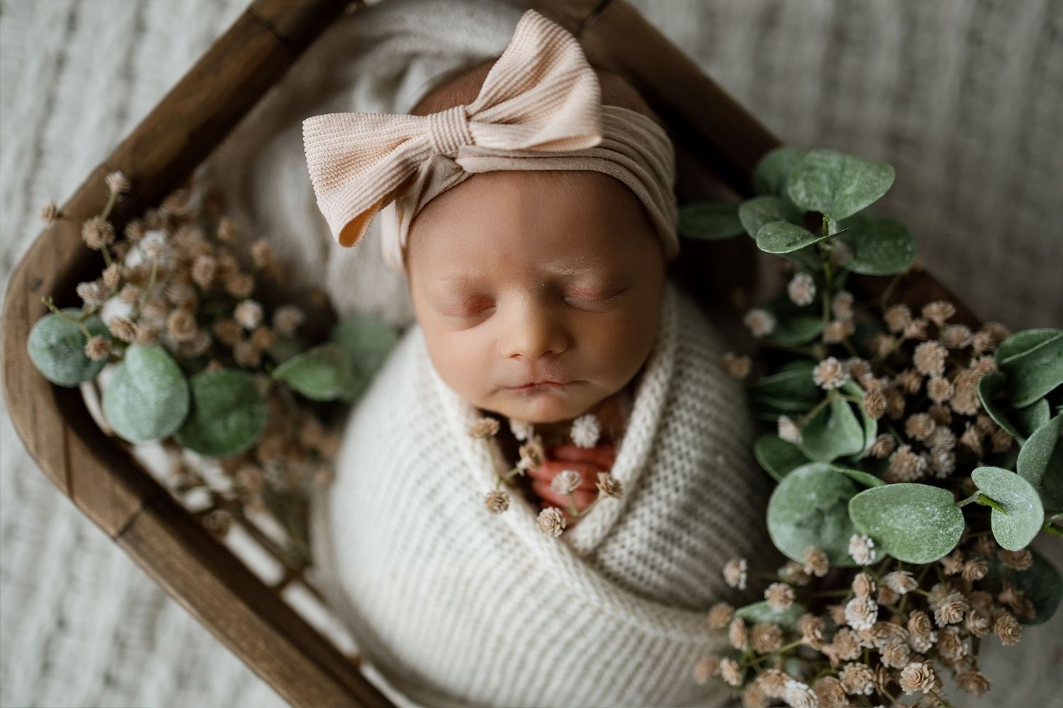 A cozy newborn photography setup with white knit blankets, dried flowers, and eucalyptus leaves in wooden boxes.