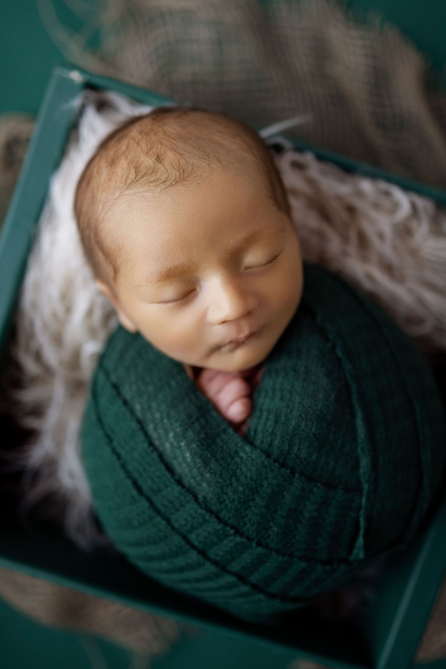Peaceful sleeping newborn swaddled in dark green wrap against teal backdrop in professional portrait series.