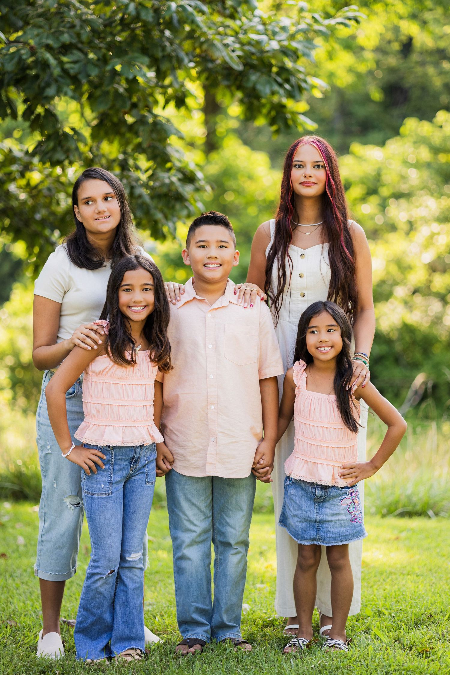 Three smiling children stand together on a grassy lawn in coordinating pink and white outfits on a sunny day.