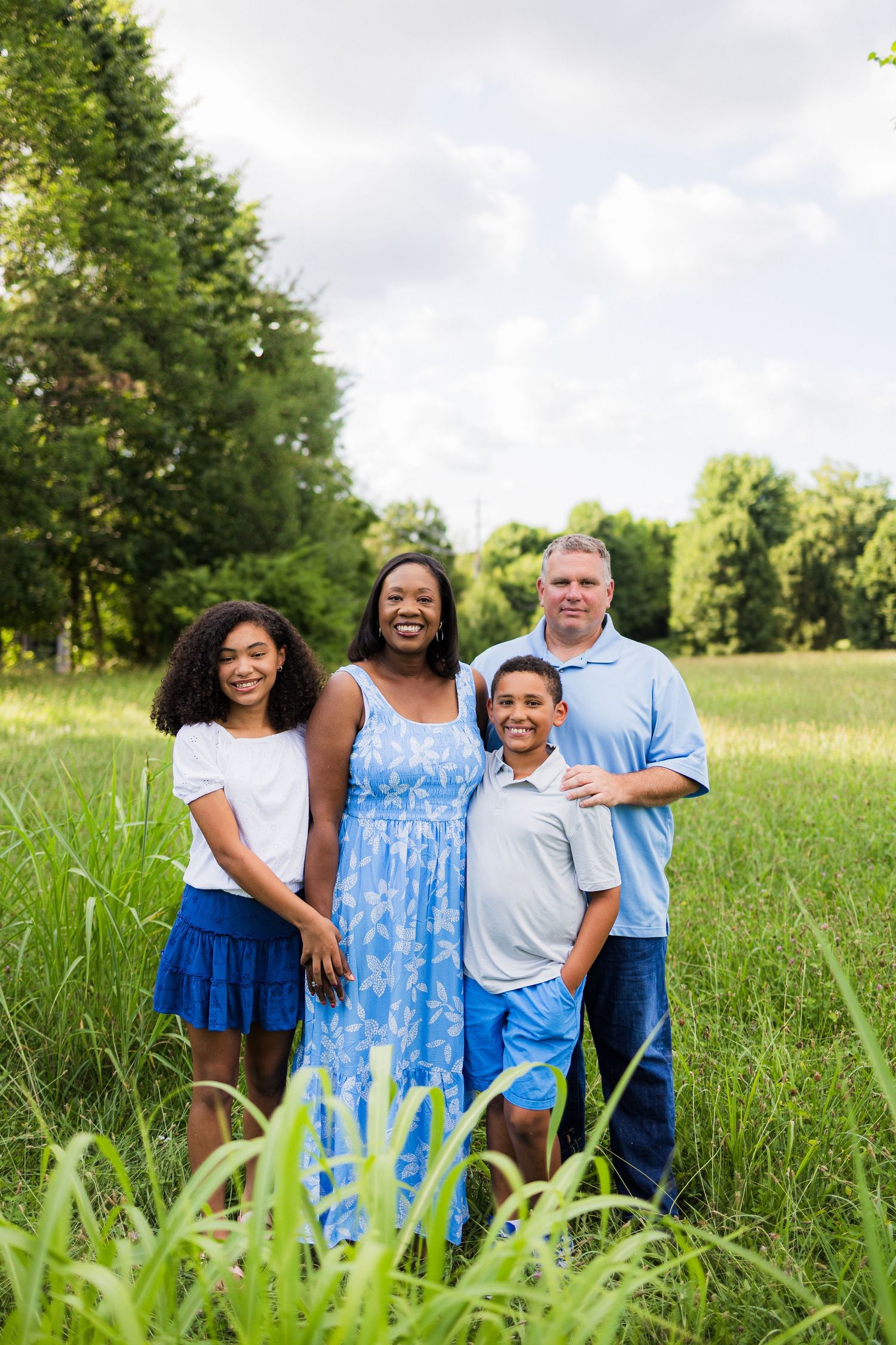 Family of four posing together outdoors in a scenic meadow with lush green trees in background.