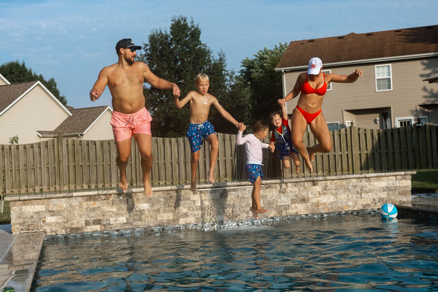 Family of five joyfully jumping into a backyard pool together on a sunny summer day.