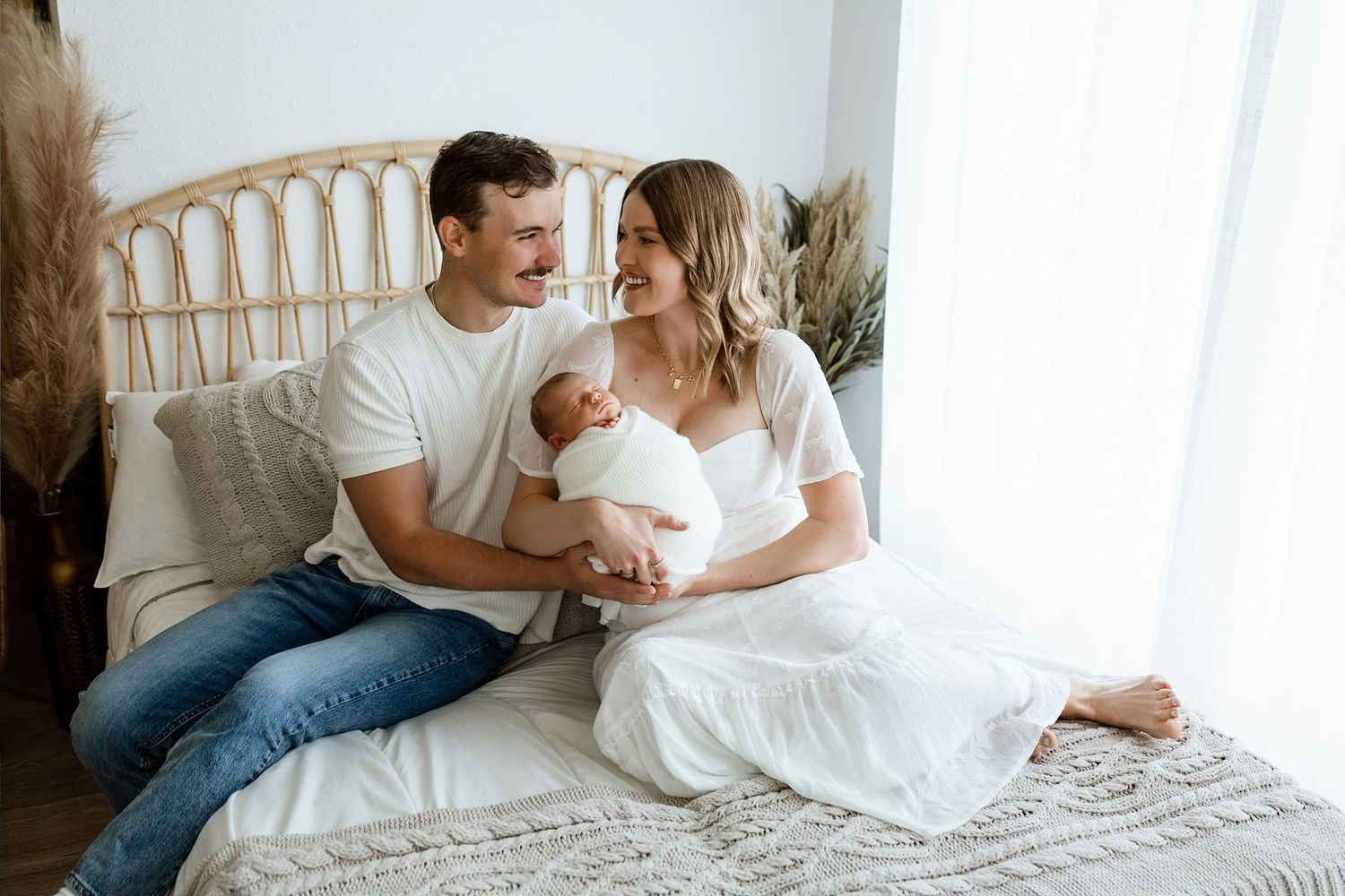 A couple sits on a bed smiling at their newborn baby wrapped in white, in a bright, cozy bedroom.