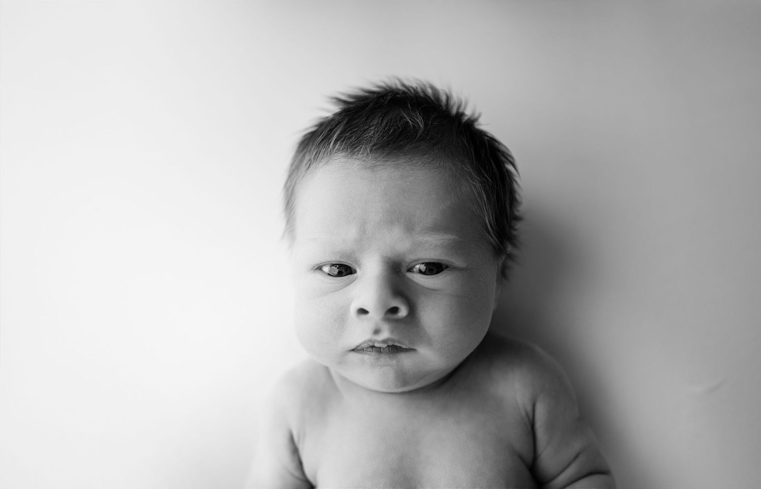 Black and white portrait of a newborn baby with furrowed brows and pouty lips lying on white background.