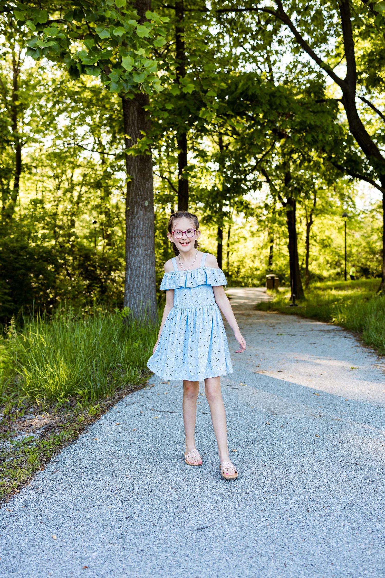 A person in a blue ruffled dress poses on a sunlit gravel path through a wooded area.